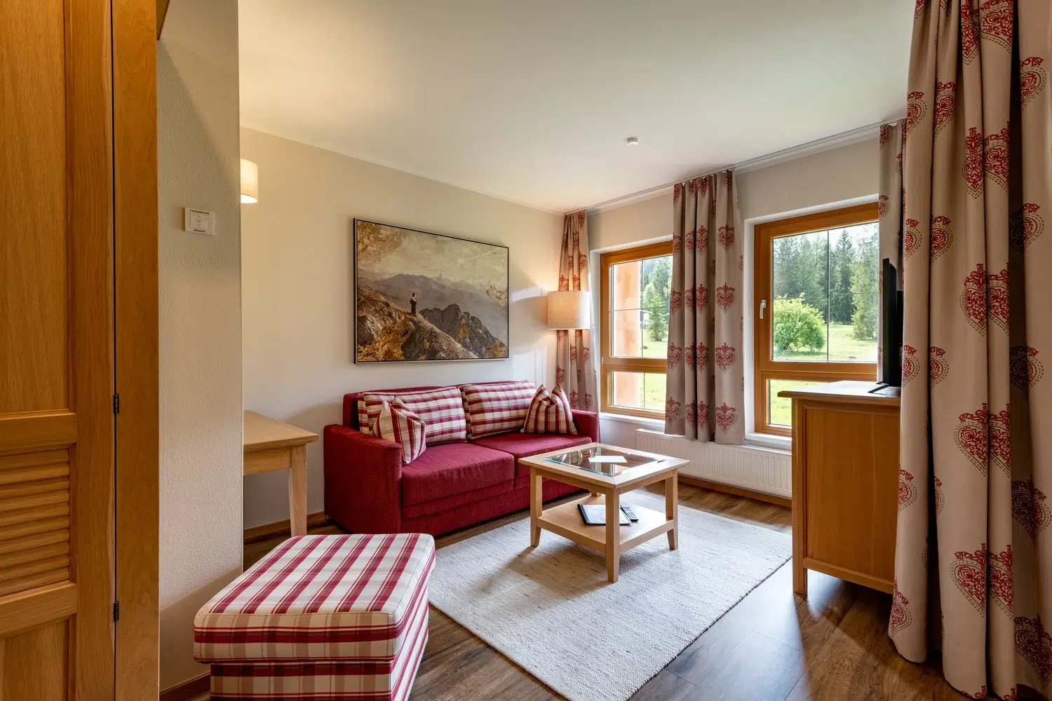 Gemütliches Wohnzimmer mit rotem Sofa, karierter Ottomane, großen Fenstern und Blick in die Natur.