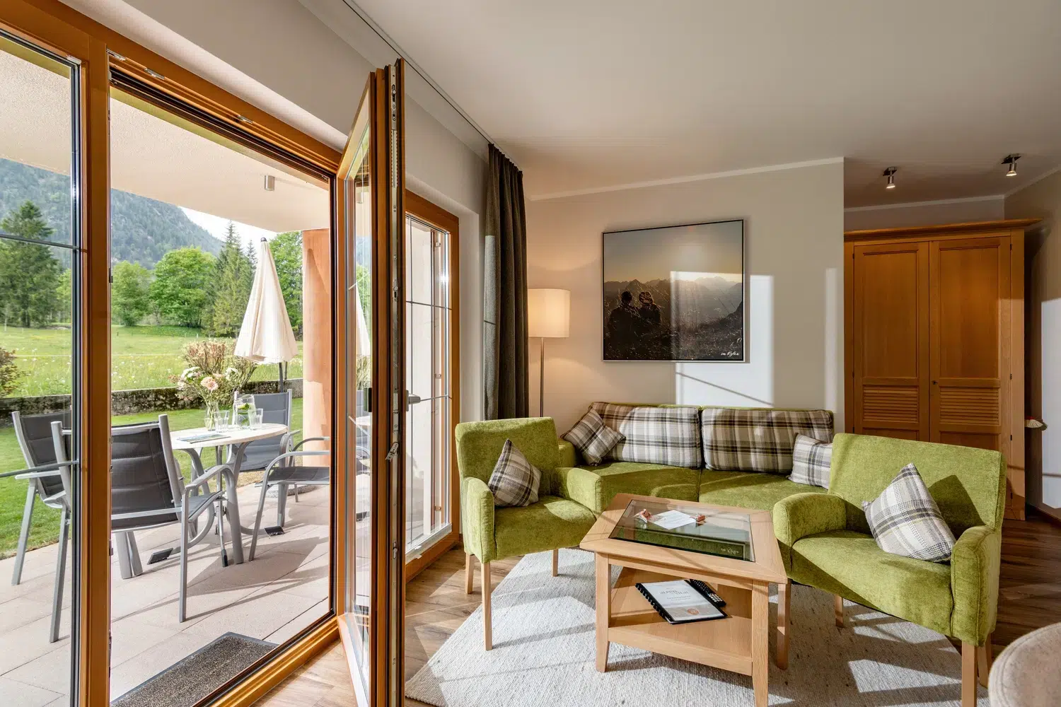 Helles Wohnzimmer mit grünem Sofa, offenen Türen zur Terrasse und Blick auf den Garten und die Berge.