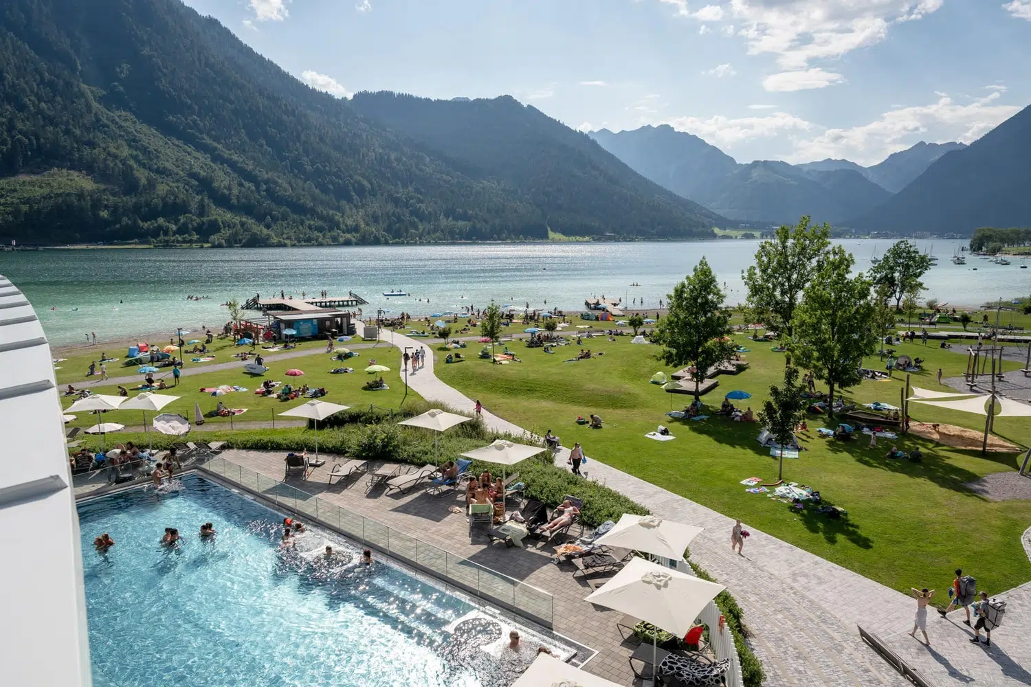 Menschen, die sich an einem Pool am See und auf einer Wiese entspannen, umgeben von Bergen und einem klaren Himmel.